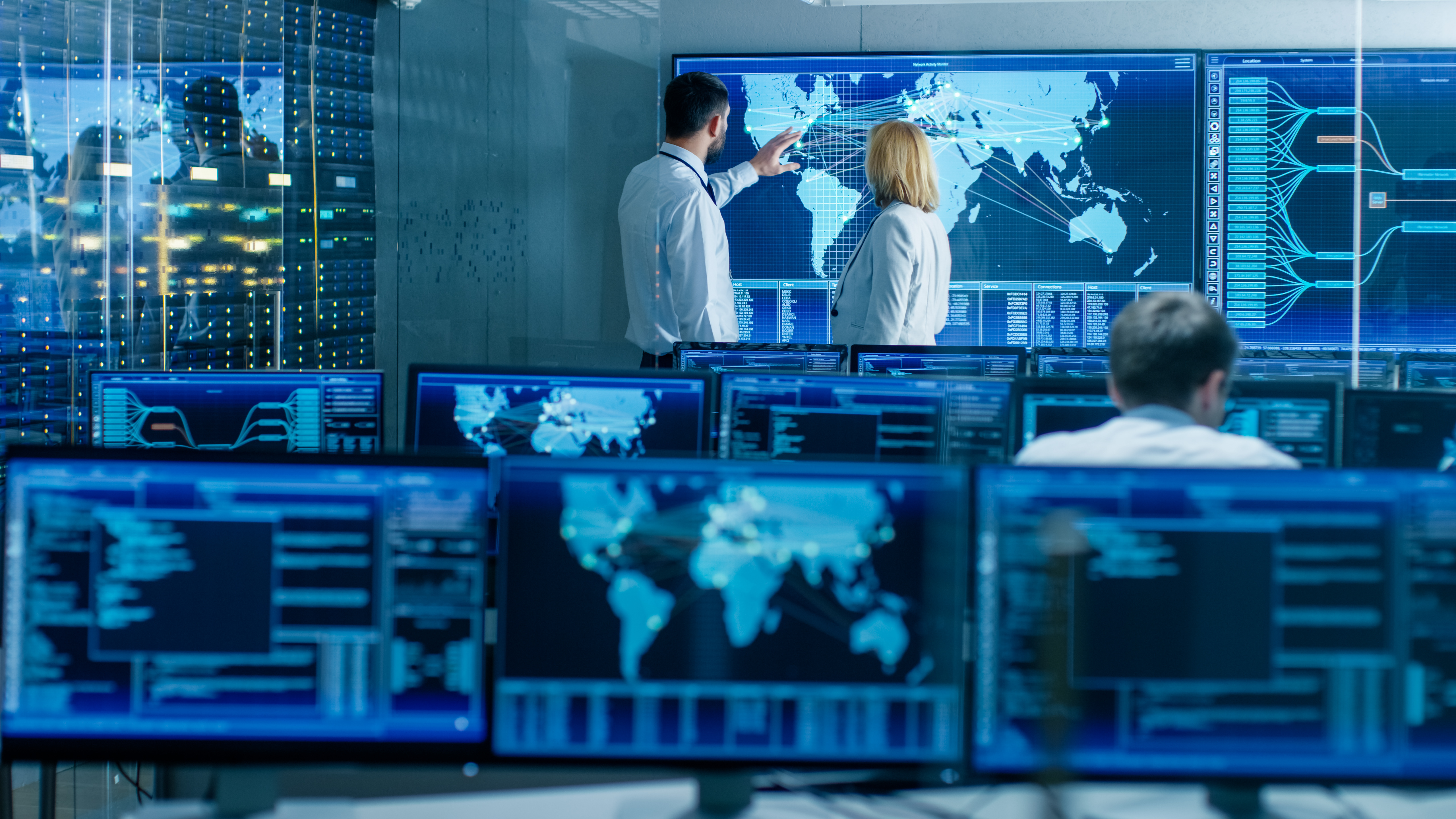 Security Operations Center (SOC) Raum mit vielen Monitore, eine Person sitztend und zwei weitere Personen stehend