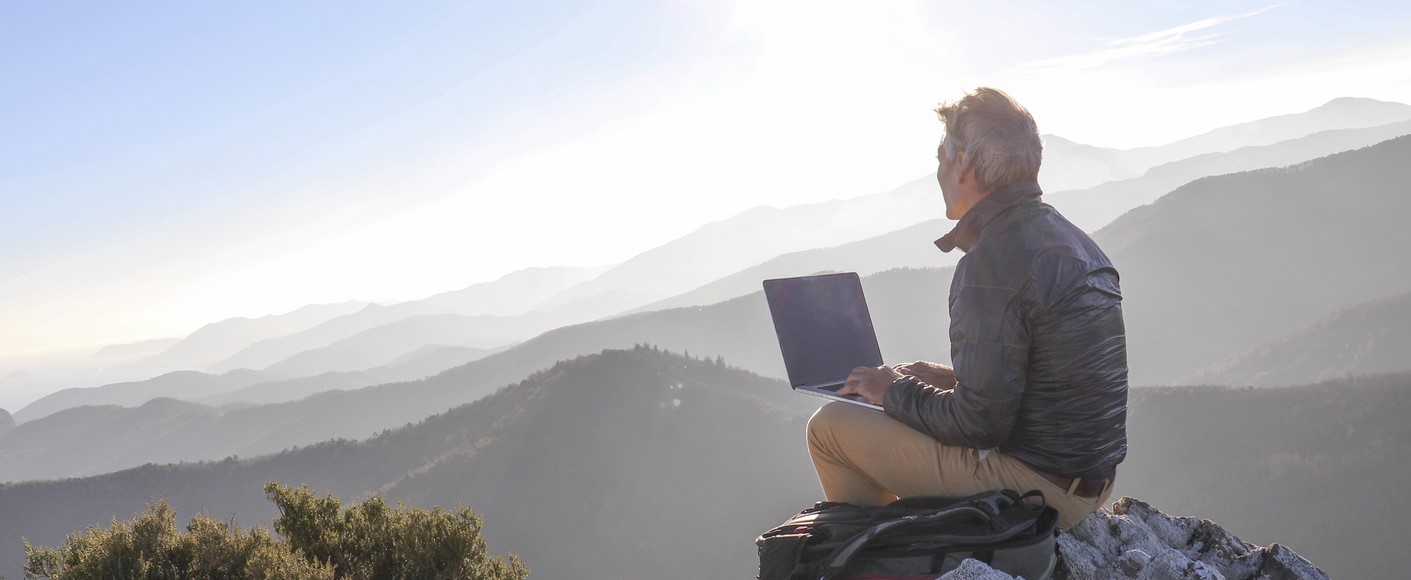 Ein Mann sitzt mit einem Laptop in einer Gebirgslandschaft und nutzt Outdoor WLAN